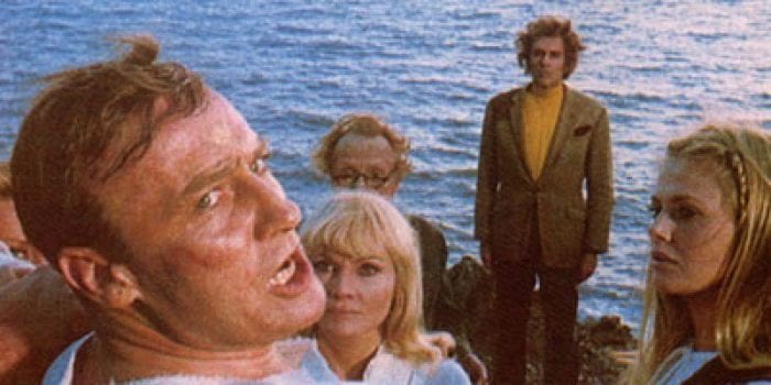 In the immediate foreground, a man's face, shiny with sweat, looks up fearfully at something behind him; in the background, a group of men and women look on, a body of water visible behind them.