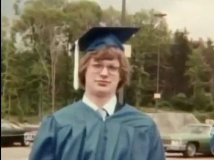 A teenage boy stands in front of a wooded area in daylight. He wears a blue graduation cap and gown.