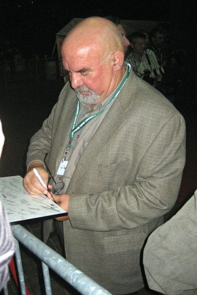 Stuart Gordon wears a laniard and signs his autograph at a convention.