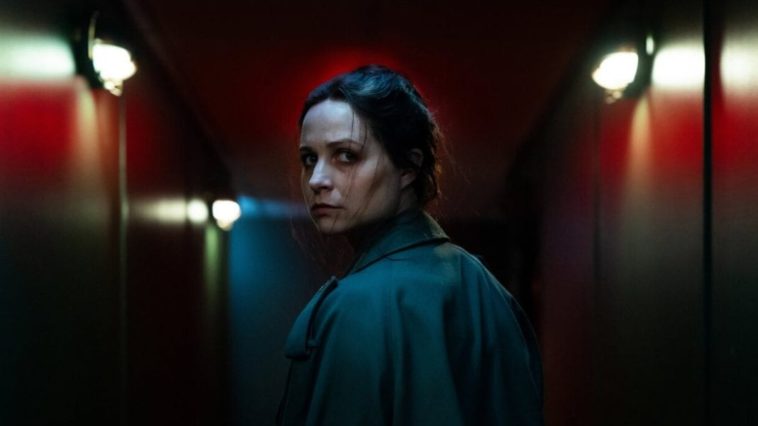Enid looks back over her left shoulder walking down a well-lit corridor