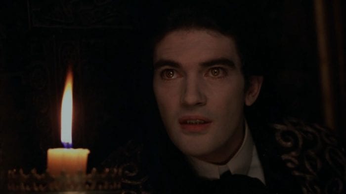 A close-up of a pale man with intense eyes sits in front of a burning candle.