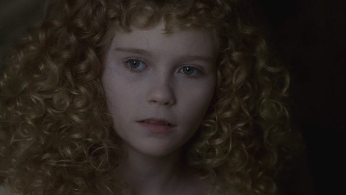 A little girl with a thick mass of golden blonde curls and extremely pale face looks into the camera. 