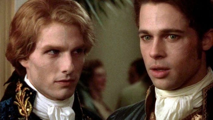 Two young men stand close young men, appearing in 18th century style hair and dress. One looks off in the distance as the other looks intensely at him as if about to say something.