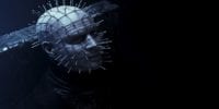 Pinhead (Paul T. Taylor), a pale man with nails sticking out of every angle of his face, sitting in a dark room. His face is somewhat illuminated by a ghostly blue light.