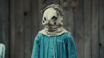 A little boy wearing a sack mask with a face drawn on it and brown fabric hair and a blue uniform looks off-camera in front of a dilapidated barn.