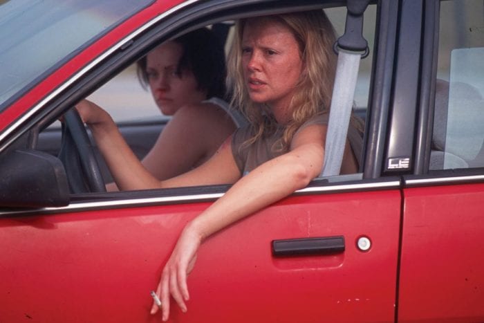 Two women sit in a red car. The driver looks disheveled and angry. The woman in the background appears frightened. 