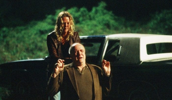 A disheveled and angry blonde woman stands behind a frightened gray haired man man crouched on his knees in front of her. She appears to be holding a gun. The area around them is dark and secluded and wooded. A car is parked in the background.