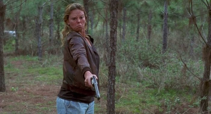 An angry, red-faced blonde woman stands in a secluded wooded area pointing a gun at an unseen target.