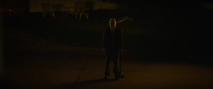 The Man in the Mask (Damian Maffei), a man wearing a burlap sack mask with two holes cut out for eyes and a drawn-on mouth, standing outside at night on what appears to be a driveway. He is dressed darkly and an axe dangles from his hand on his left side.