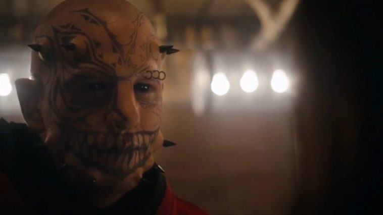 The Devil (Damian Maffei), a pale man with pitch black eyes in a room lit by a few exposed lightbulbs. He has tattoos on his face, most notably a large, sharp-toothed smile across his mouth. He has three black spike implants on either side of his jawline, and two on each of his temples.