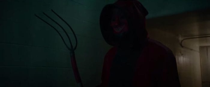 The Devil (Damian Maffei), a figure in a red hood wearing a red devil mask, standing in dim hallway. He is holding a three-pronged rake that resembles a pitchfork.
