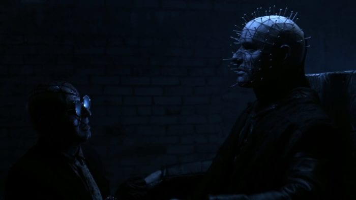 The Auditor (Gary J. Tunnicliffe), a man with slash scars all across his head and face wearing a pair of round, dark sunglasses which are reflecting a blue light in front of him. He is kneeling in front of Pinhead (Paul T. Taylor), a ghostly pale man with pins sticking out of almost every angle of his face. He is sitting in some sort of throne-like chair, and is partially illuminated by that soft blue light behind him.