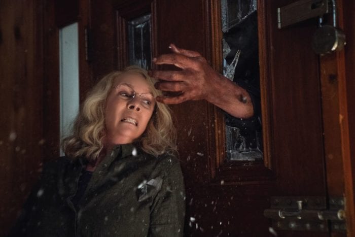 Jamie Lee Curtis as Laurie Strode, her back against a door, looking in terror as the hand of Michael Myers bursts through the door's glass panel.