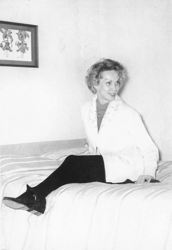 Black and white photograph of Maila Nurmi without makeup sitting on a bed and smiling at someone outside of the frame.