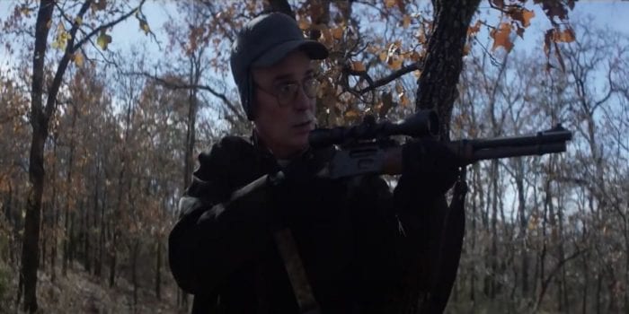 Paul T. Taylor as Frank Hughes in Whitetail, an older pale man wearing glasses and a cap covering his ears. He is in the woods, dressed warmly and aiming a shotgun at something in front of him. He looks a bit apprehensive.