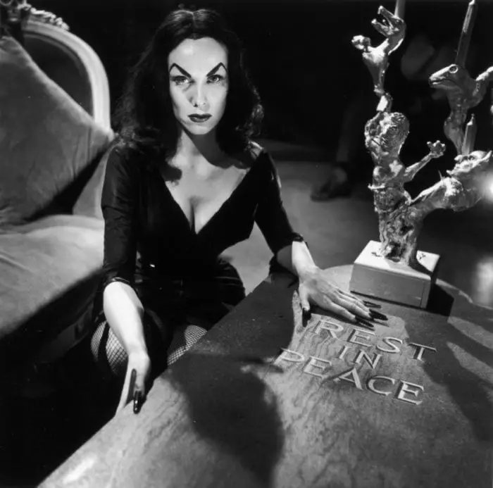 Maila Nurmi as Vampira sits nexzt to a coffin that says "Rest In Peace" on The Vampira Show.