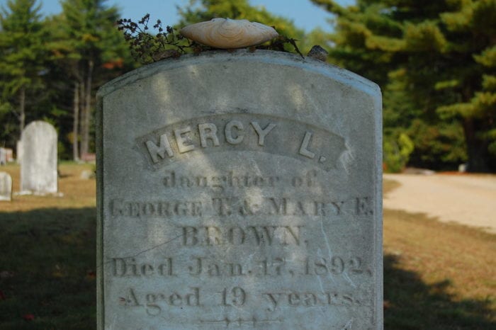 A day time shot, a close up of an old, worn headstone in a graveyard. The name Mercy L. Is inscribed in the grey stone. 