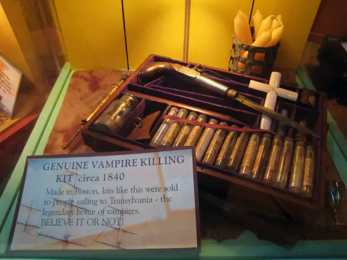 A brown wooden box with wooden stakes and a large wooden cross sits on a table with a sign next to it that reads "Genuine Vampire Killing Kit., Circa 1840."