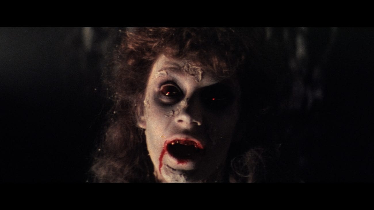 A pale feminine face with long curly dark hair peers out of the darkness with large dark eyes and blood dripping from her mouth.