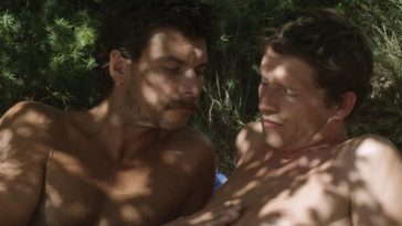 Two shirtless men on a beach in the shade of a tree, with one caressing the other's chest.