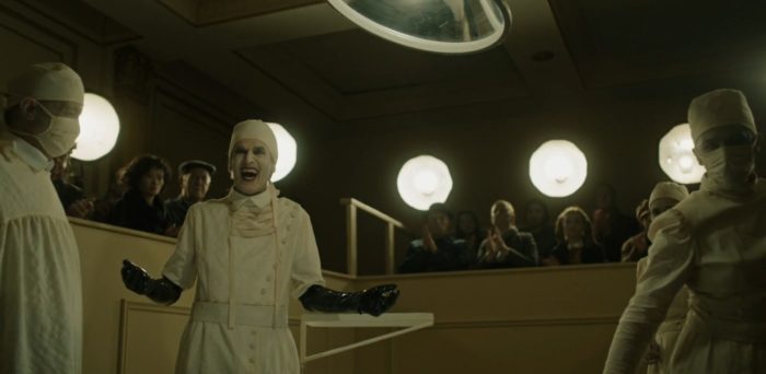 A surgeon in white makeup laughs in a surgery theater complete with an audience
