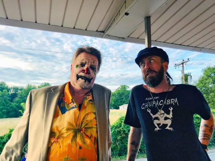 Behind the scenes photo of Kill Giggles with actor Vernon Wells (a husky older white man with gray hair wearing a light orange Hawaiian shirt) in his Giggles character makeup (patchy white paint, deep red nose, deep blue lips and bright green splotches) and director Jaysen Buterin (a tan, bearded man in a black had and a black t-shirt that reads "save the Chupacabra" with an illustration of said cryptid underneath). The two are standing underneath a roof, behind them a sunny day, the sky littered with clouds and the trees glowing bright in the sunshine.