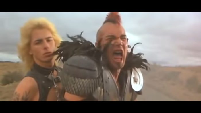 Vernon Wells as Wez in Mad Max 2, a white man with a red and black mohawk and armor. His shoulder pads are adorned with black feathers. He is on some sort of vehicle unseen, and is shouting at someone out of frame. Sitting behind him is another white man with fluffy blonde hair and a leather garment. They are outside on a long road stretching through a desert-like place. 