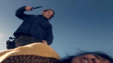 Nick Stahl in Fear The Walking Dead as Riley, a white man with long-ish brown hair wearing a dark blue coat and jeans. The camera is pointed upwards at him, a woman on the ground blurred at the base of the shot, as Riley stands over her, backdropped by the blue sky. He is wielding a knife in his right hand, poised to attack.