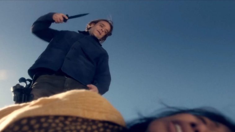 Nick Stahl in Fear The Walking Dead as Riley, a white man with long-ish brown hair wearing a dark blue coat and jeans. The camera is pointed upwards at him, a woman on the ground blurred at the base of the shot, as Riley stands over her, backdropped by the blue sky. He is wielding a knife in his right hand, poised to attack.