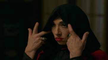 Naomi Grossman in American Horror Story: Apocalypse as Samantha Crowe, a white woman with red lips and dark eye makeup wearing a black hoot over her head. Her hands are raised to pull the hood back.