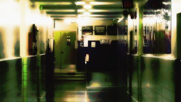 A badly lit, decrepit school hallway is barren of life