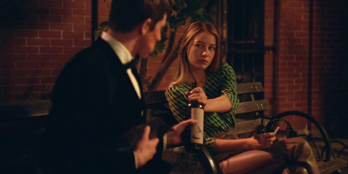 Lena sits on a park bench leaning over toward the camera to hand Richard a bottle of wine.
