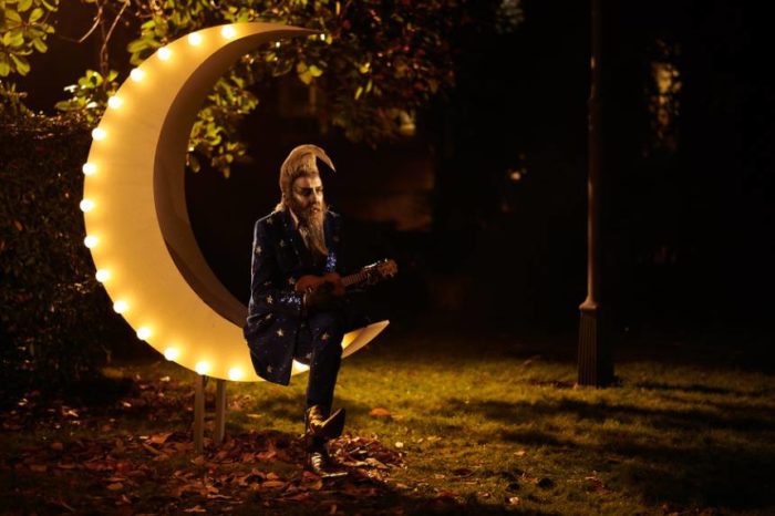 Frank Metterton sits on a crescent moon covered in lights playing a ukulele 