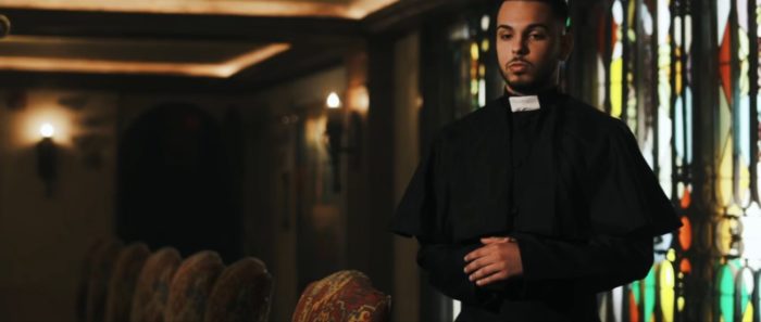 Joey Limongelli as Father Leo (a brown-skinned man with soft black facial hair and backswept black hair) in a room in a church, illuminated by soft yellow lights and sunlight filtering in through the stained glass behind him. He is wearing black robes and his arms are folded gently across his middle.
