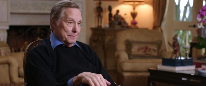 William Friedkin, now in his mid-80s, sitting, wearing a dark colored sweater over a collared shirt; slightly out of focus in the background are stuffed chairs, a lamp, and tables on which sit various figurines. 