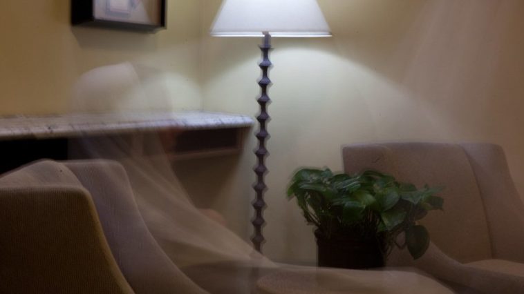 A translucent hooded figure dressed in white sits in a chair in a living room.