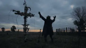 Shadow raises his arms to the sky standing in front of a cross decorated with animal skulls