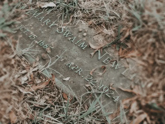 A headstone on the ground is covered with grass and leaves.
