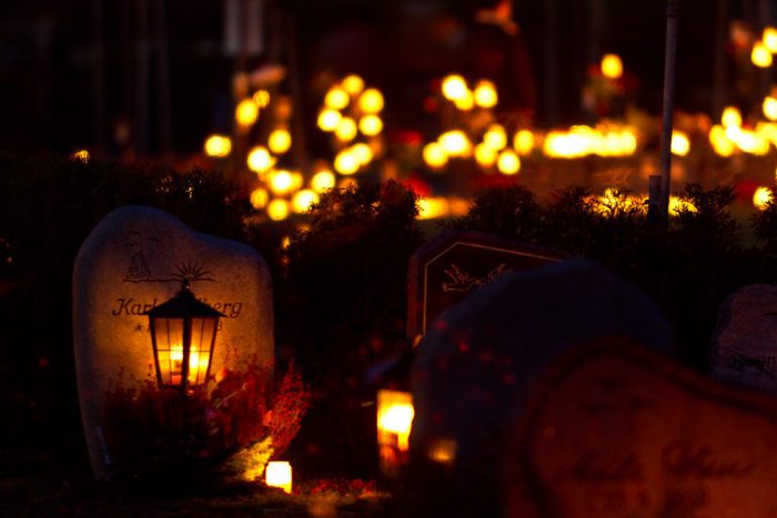 A close shot of headstones in a cemetery at night. Small, glowing lanterns hang from them..