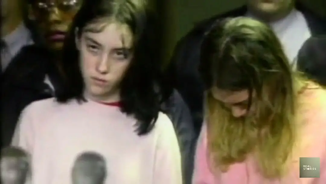 A picture of two teen girls. A dark haired girl looks up to the camera with a very unhappy expression. The other looks down, her blonde hair covers her face.