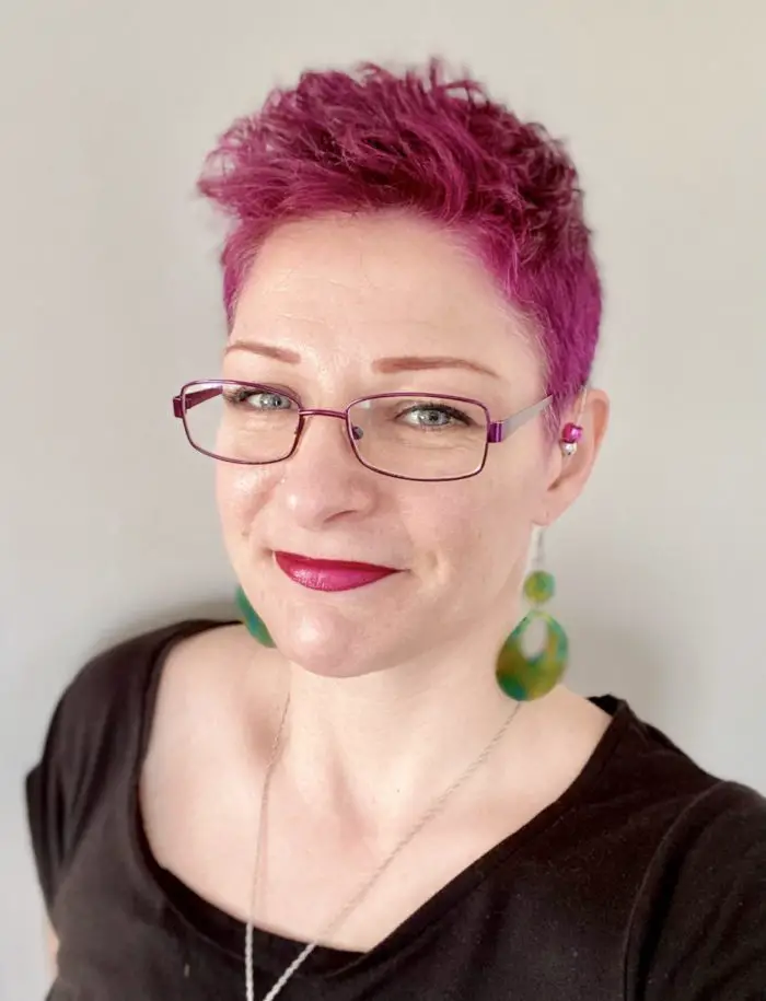 A portrait photo of Tabatha Wood, author. They have red-rimmed glasses and short pink hair.