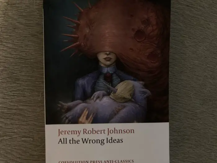 Cover for All the Wrong Ideas has a woman holding a bizarre doll wearing a mask while she's consumed by a slug-like creature