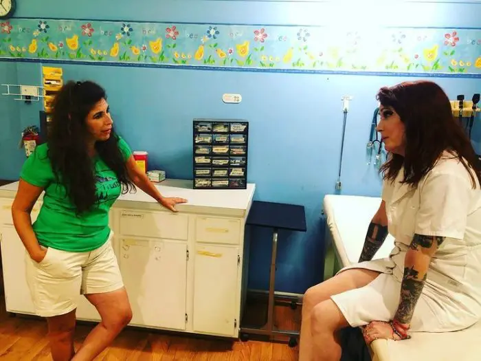 Felissa Rose (a tan woman with fluffy dark brown hair wearing a green t-shirt and white shorts) in a doctor's office, blue with yellow chicks and blue and red flowers painted on the wall, with Julie Anne Prescott (a pale woman with tattooed arms, deep red hair and heavy eyeshadow wearing a white button-up gown). Felissa is leaning casually on a desk, one hand in her pocket.