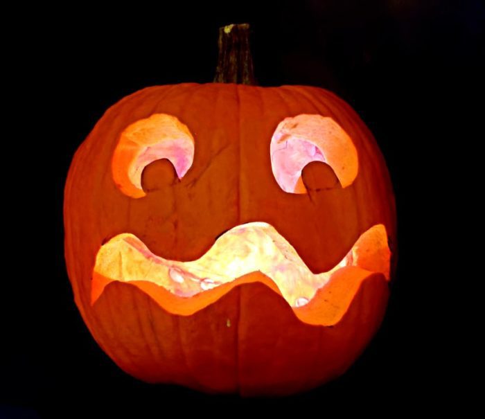 Wobbly mouthed screaming pumpkin carved in a jack-o'-lantern, lit by a candle inside, glowing in the dark