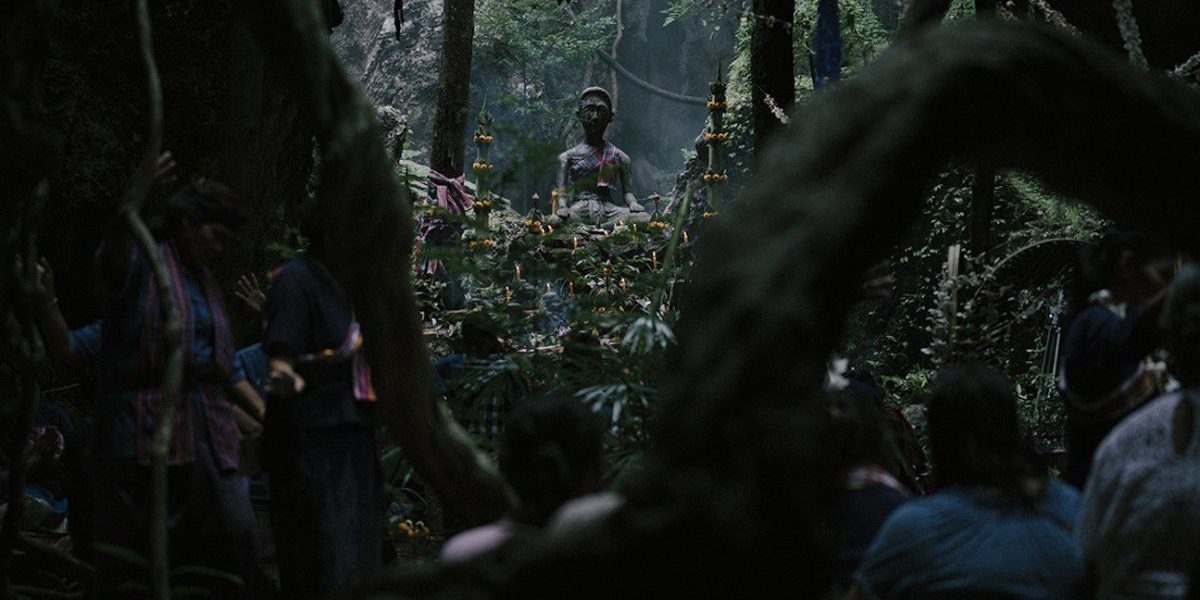 A statue of Ba Yan, a Thai deity, sits in the woods surrounded by offerings of flowers and food
