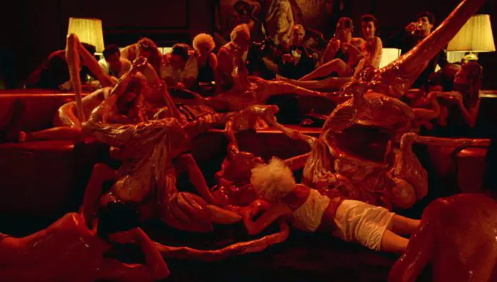 A group of men and women sit and lay around a room, their bodies melting together, covered in sweat and slime
