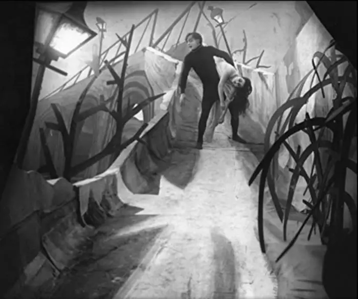 The main character in Cabinet of Dr. Caligari creeps down a pathway carrying an unconscious female. The path is angled oddly, a good example of German Expressionism.