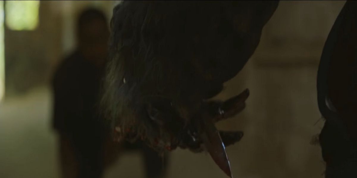 A large, gray-furred monster's hand is in focus on the screen against a dim, yellow-toned location and a man kneeling in front of the beast. Its long claws are splattered with blood.
