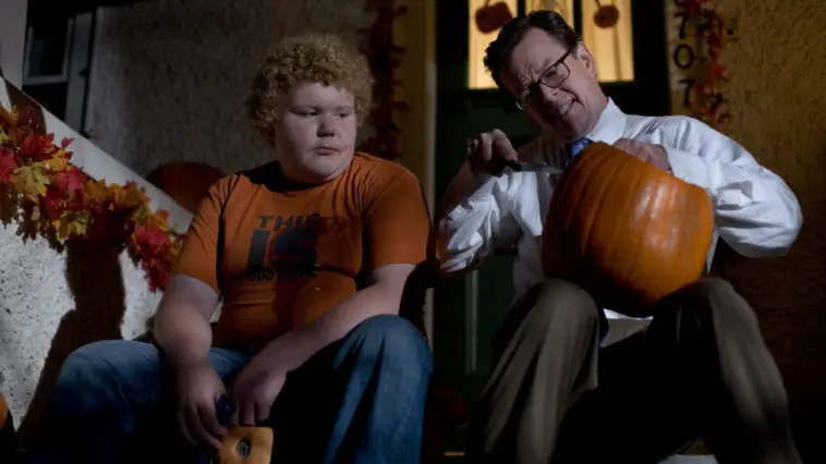 A kid sitting on the porch with a man carving a pumpkin