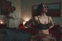 a woman sits on a bed while a man buttons his shirt in the background